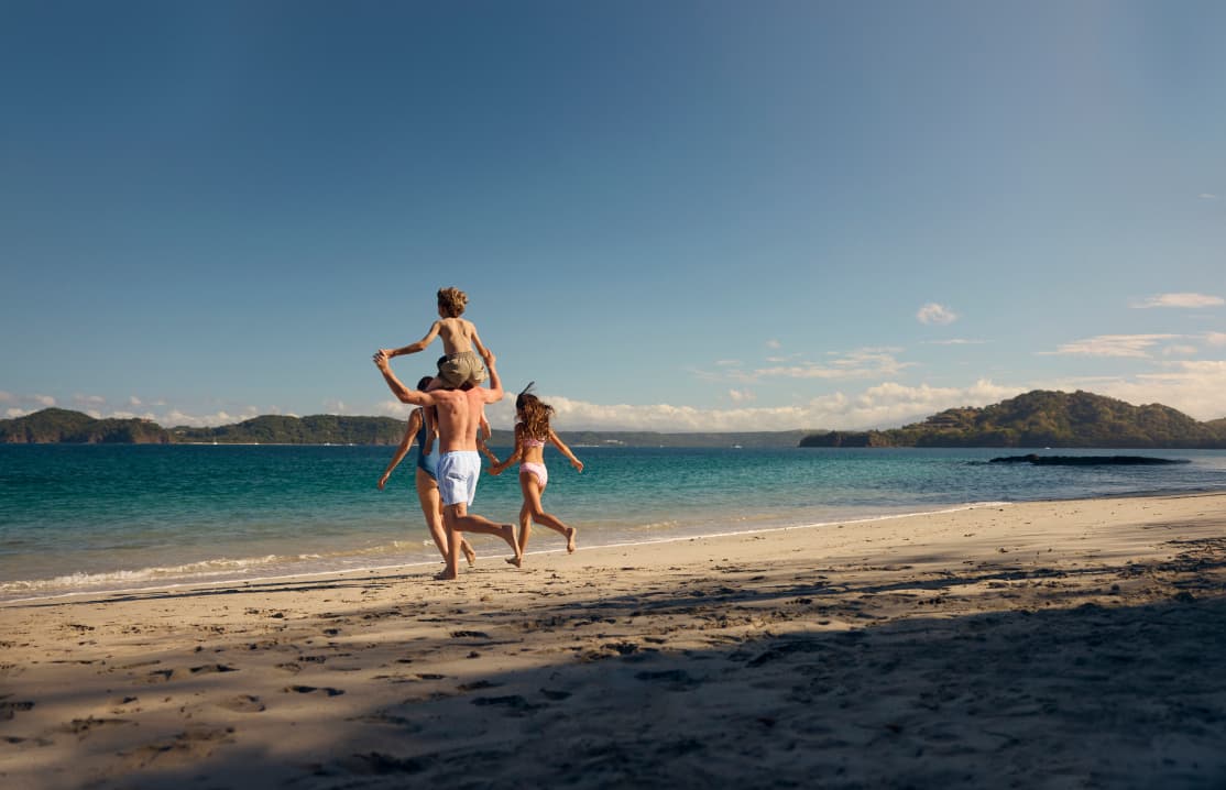 Quiet, sandy shores of Playa Penca, Costa Rica with calm, turquoise Gulf of Papagayo, rocky outcrops along the shoreline, and tropical vegetation surrounding the cove. A family of 4 is walking towards the water.