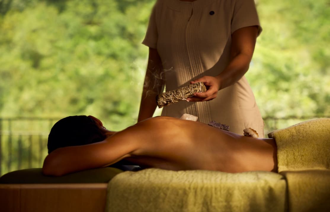 Serene spa treatment room with a massage table, soft lighting, natural wood finishes, and a large window opening to lush tropical vegetation.