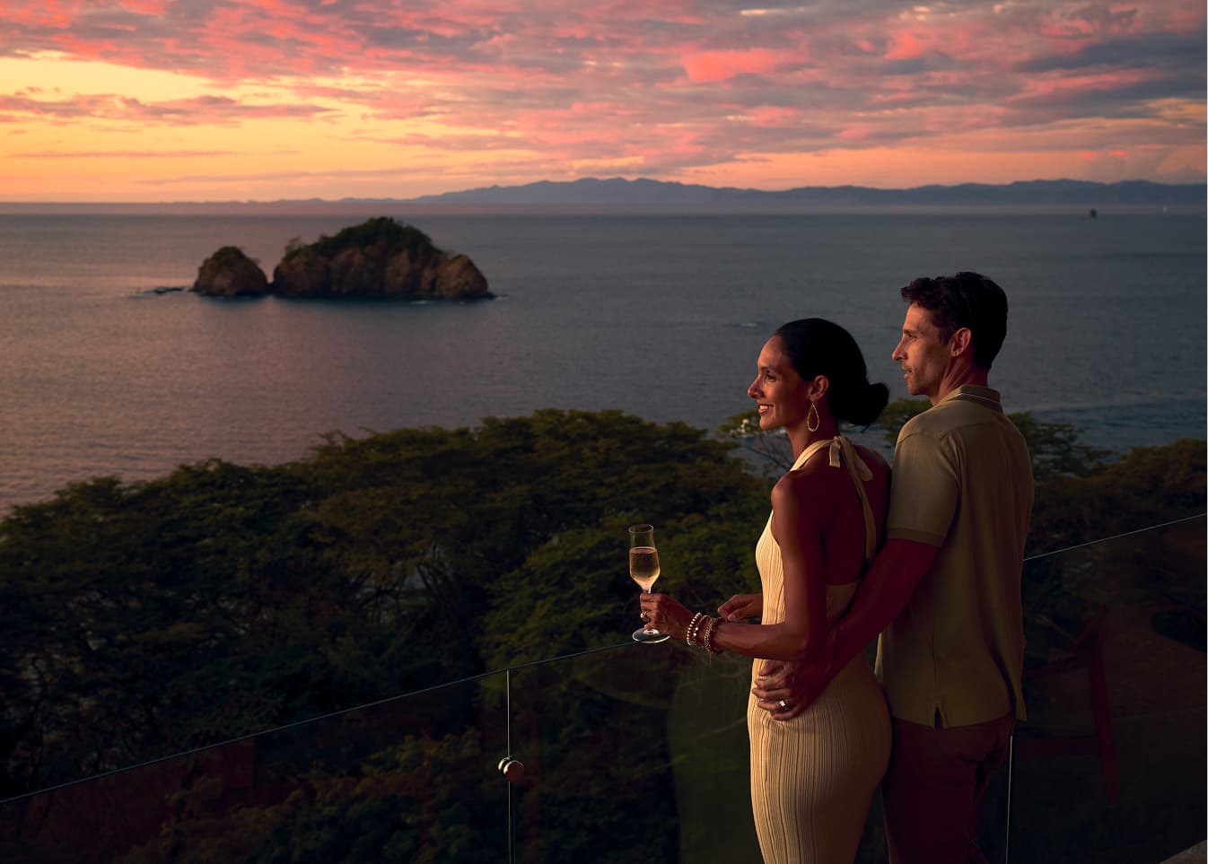 Couple standing on a cliffside terrace overlooking the Gulf of Papagayo, with their arms around each other and the sun low over the water.