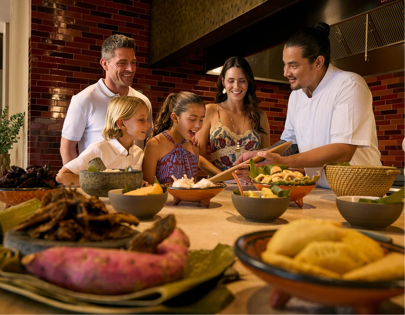 Chef preparing food for a smiling family of four in an open kitchen, with bowls of fresh ingredients on the countertop, illustrating a personalized in-home dining experience.