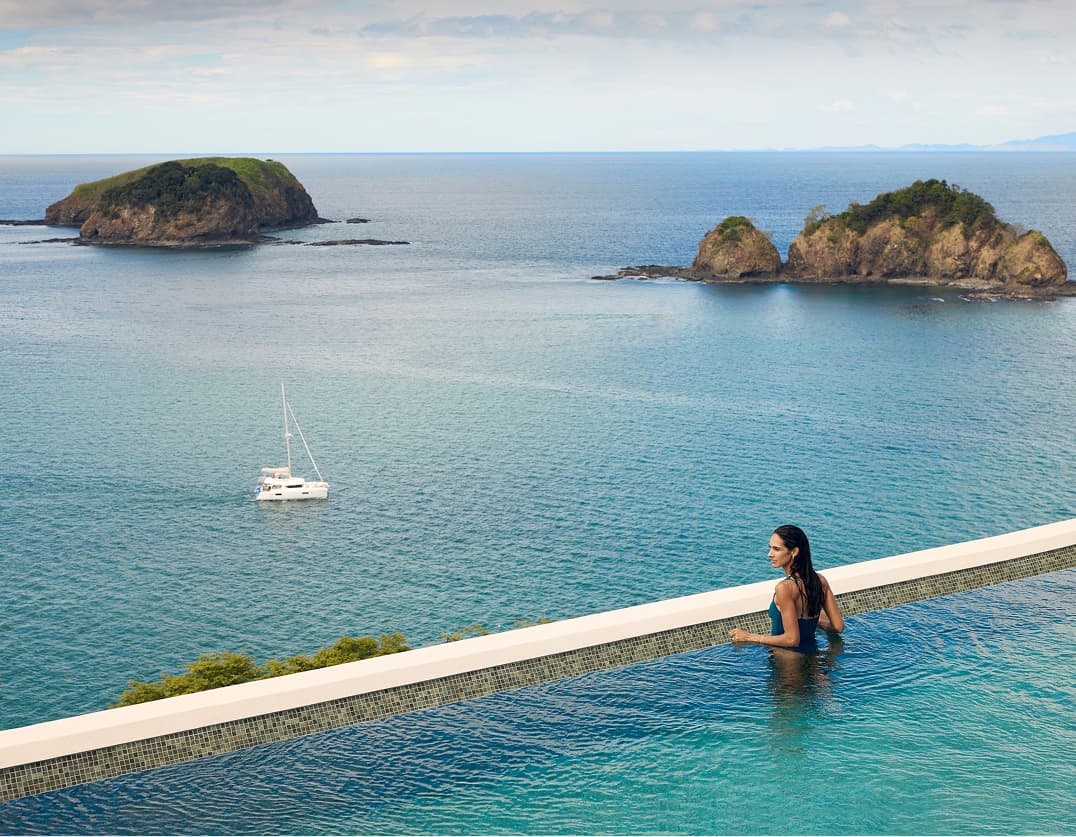 Person in an infinity pool overlooking the ocean with rocky islands and a sailing boat in the water beyond, capturing a tranquil seaside resort setting.