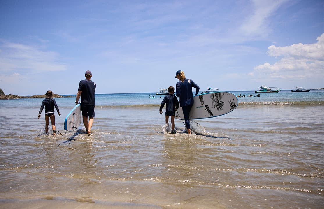 family walking into gentle ocean waves holding surfboards on a bright sunny day