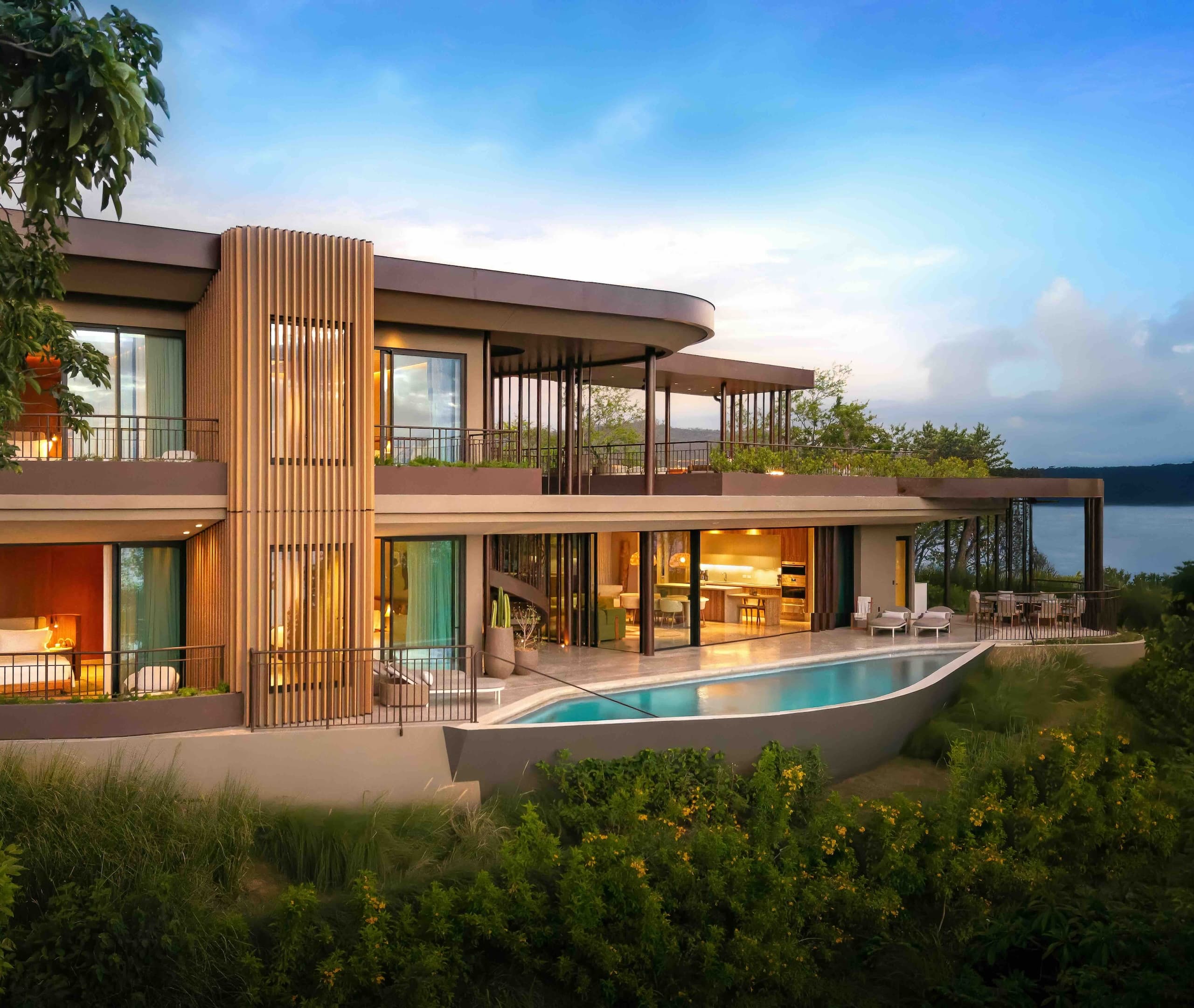 Exterior of a luxury estate home at the Waldorf Astoria Punta Cacique with large windows, multiple levels, outdoor seating areas, and tropical landscaping on a hillside overlooking the ocean.